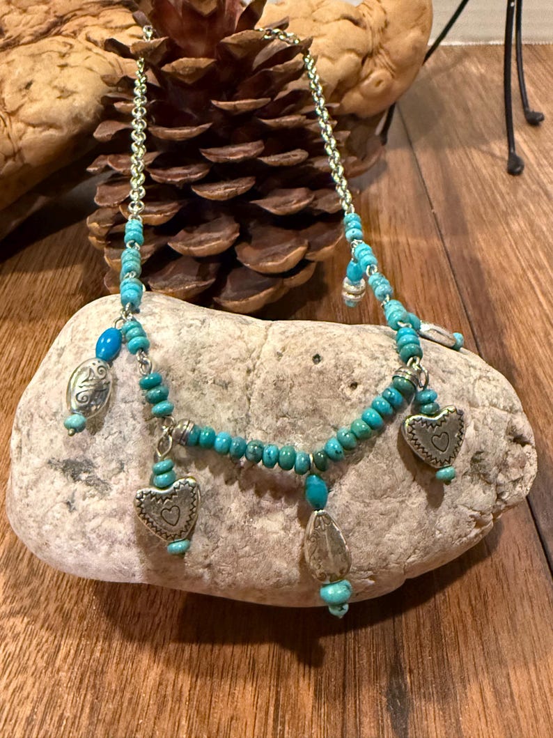 May include: A turquoise beaded necklace with silver-toned heart and teardrop charms. The necklace is displayed on a light-colored rock against a wooden background. The necklace has a chain and turquoise beads.