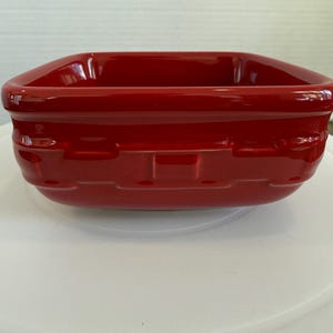 May include: A square, red ceramic basket-style dish. The dish has a glossy finish and a woven basket design on the sides. The dish is sitting on a white surface.