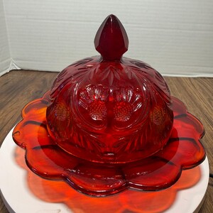 May include: A vibrant red glass butter dish featuring a domed lid with a pointed finial and embossed floral patterns. The base has a scalloped edge, and the entire piece is crafted from translucent, deep red pressed glass with a glossy finish.