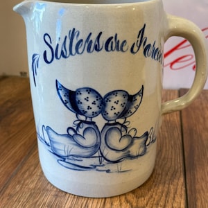 May include: A beige ceramic jug with a spout and handle. The jug features blue illustrations of two figures in bonnets and the words "Sisters are Forever". The design is hand-painted.