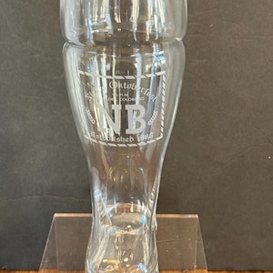 May include: Clear glass beer stein with a flared base and a wide rim. The stein has the words "Oktoberfest" and "NB Established 1985" etched on the front. The stein is displayed on a clear plastic stand.