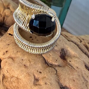 May include: A handcrafted ring featuring a large, faceted black gemstone set in a gold-toned wire wrap design. The ring has a unique, artistic style, with a coiled band and intricate wirework around the stone. The ring is displayed on a piece of driftwood.