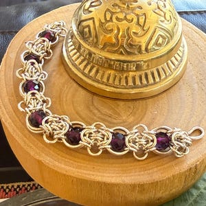 Op de afbeelding: Een zilverkleurige chainmail armband met donkerpaarse kralen. De armband wordt getoond op een houten oppervlak naast een goudkleurige bel. De bel heeft ingewikkelde patronen en een afgeronde bovenkant.
