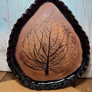 May include: A teardrop-shaped ceramic dish featuring a dark brown, ruffled edge and a textured, light brown center. The interior surface displays a detailed, dark brown embossed leaf pattern with visible veins, creating a natural aesthetic.