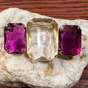 May include: A gold-toned brooch with three faceted glass stones. The center stone is clear and the two outer stones are a deep purple.