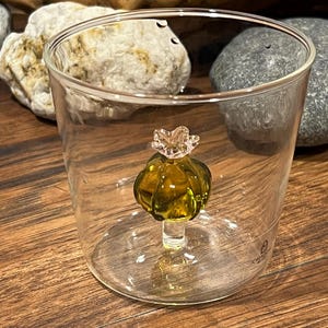 May include: A clear glass cup with a green glass poppy flower inside. The poppy has a pink center and a green stem.