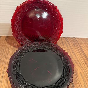 May include: Two ruby red glass plates with ornate, scalloped edges. The plates have a detailed embossed pattern around the rim and a smooth center. One plate is positioned above the other, showcasing the rich color and design.