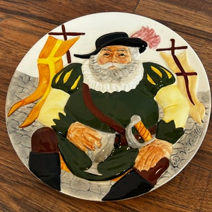 May include: A ceramic plate with a raised image of a man in a green and yellow costume holding a sword. The plate has a white background with a brown border and the text "D.V. Toole" in the lower right corner.