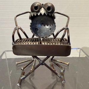May include: A whimsical metal sculpture of a chair with a creature-like face. The chair features a grill-like seat, spindly legs, and a face with large, round eyes and a jagged mouth. The sculpture is made of welded metal.