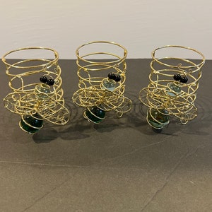 May include: Three gold wire candle holders with green and black glass beads. The holders are in the shape of a cylinder with a flared base.
