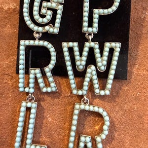 Op de afbeelding: Een paar statement oorbellen die het woord "GIRL POWER" spellen in zilver metaal. Elke letter is omrand met kleine, lichtblauwe kralen. De oorbellen hangen aan een zilveren haak en worden gepresenteerd tegen een zwarte achtergrond.