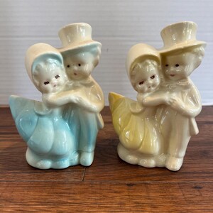 May include: A pair of vintage ceramic figurines of a couple holding each other. The figurines are painted in pastel colors, with the woman in blue and the man in yellow. They are both wearing hats and have a sweet, innocent expression on their faces.
