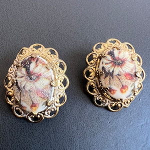 May include: A pair of gold-tone earrings with a floral design. The earrings are oval shaped and have a delicate filigree design around the edge. The flowers are white with red and brown accents.