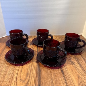 May include: A set of six ruby red glass teacups and saucers. Each cup has a handle and sits on a matching saucer. The cups and saucers have a detailed, embossed pattern. The set is displayed on a wooden surface.