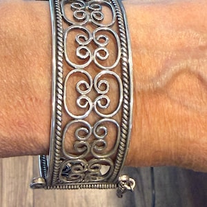 May include: Silver cuff bracelet with an openwork design of swirling patterns. The bracelet has a hinged closure and a textured border. This statement jewellery piece is approximately 5 cm wide.