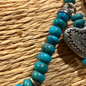May include: Close-up of a turquoise beaded necklace with a silver heart pendant. The necklace features round, polished turquoise beads and a decorative silver heart charm with a detailed edge. The background is a woven, textured surface.