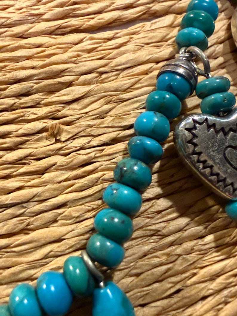 May include: Close-up of a turquoise beaded necklace with a silver heart pendant. The necklace features round, polished turquoise beads and a decorative silver heart charm with a detailed edge. The background is a woven, textured surface.