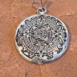 May include: A silver-colored pendant with a detailed, circular design. The pendant features an intricate pattern of symbols and shapes, possibly representing a calendar or celestial map. The pendant has a loop at the top for a chain.