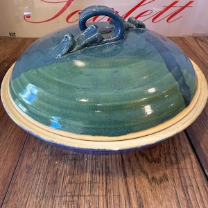 May include: A handcrafted ceramic serving dish with a lid. The dish features a two-tone blue and green glaze with a cream-colored rim. The lid has a curved handle and decorative accents. The dish is designed for serving food.