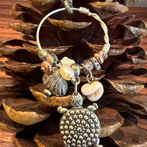 Puede incluir: Un collar de plata con un dije de tortuga de plata, un dije de concha de mar de plata y una variedad de cuentas. El collar está colgado de una piña.