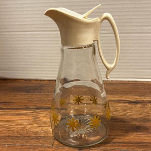 May include: A clear glass pitcher with a white plastic lid and handle. The pitcher has a gold snowflake design.