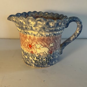 May include: A ceramic creamer with a scalloped rim and a handle. The creamer features a hand-painted design of a sheep, sun, and flowers. The body of the creamer has a blue and pink speckled pattern.