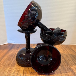 Op de afbeelding: Drie robijnrode kelkglazen met sierlijke bloemmotieven. De kelkglazen hebben een ronde voet, een lange steel en een komvormige bovenkant. De kelkglazen zijn gemaakt van glas en zijn gerangschikt op een houten oppervlak.