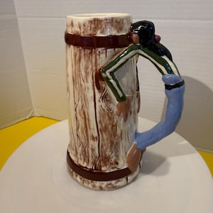 May include: A ceramic beer stein with a wood-grain design. The handle is a figure of a person wearing a green and white striped shirt and blue jeans. The mug has a brown rim and base.