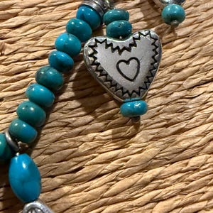 May include: Close-up of a turquoise beaded necklace with silver-toned heart and teardrop pendants. The heart pendant has a small heart cutout. The necklace is on a woven, textured surface.