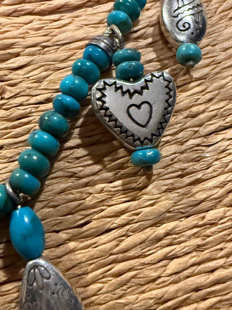 May include: Close-up of a turquoise beaded necklace with silver-toned heart and teardrop pendants. The heart pendant has a small heart cutout. The necklace is on a woven, textured surface.
