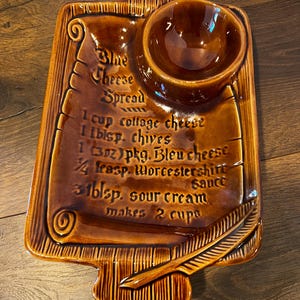 May include: Brown ceramic serving dish with a recipe for blue cheese spread inscribed on it. The dish has a small bowl for dipping and a decorative feather design.