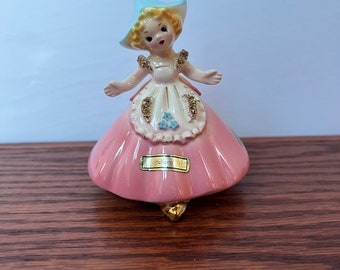 May include: A porcelain figurine of a girl wearing a pink dress, blue hat, and holding a bouquet of blue flowers. The figurine is labeled "Holland".