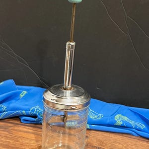 May include: A vintage glass and chrome hand-crank food chopper with a light blue wooden knob. The chopper has a clear glass jar with markings for measuring.