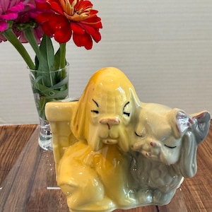 May include: A vintage ceramic planter in the shape of two dogs, one yellow and one light gray, embracing. The planter has a rectangular opening for plants. A small glass vase with red and pink flowers is in the background.