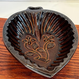 May include: A heart-shaped, brown ceramic dish with a handle. The dish features a floral design in the center and a ribbed texture around the edges. The glossy finish enhances the details of the embossed flowers and leaves.
