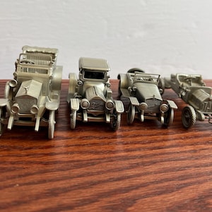 Op de afbeelding: Vier vintage-stijl tinnen modellauto's, waaronder een Rolls Royce, een Cadillac, een Mercedes en een vintage raceauto. De auto's zijn allemaal in een zilveren afwerking en worden getoond op een houten oppervlak.