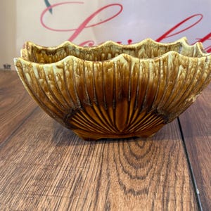 May include: A brown ceramic bowl with a scalloped rim and a textured, ribbed exterior. The bowl has a wide, open shape and sits on a wooden surface. The bowl's color transitions from a lighter brown at the rim to a darker brown at the base.