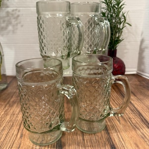 May include: Four clear glass beer mugs with diamond-patterned sides and sturdy handles. The mugs are arranged on a wooden surface, with a few sprigs of greenery in the background. The mugs are ideal for serving cold beverages.