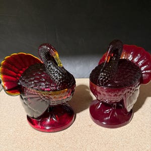 May include: Two ruby red glass turkey figurines. The turkeys have detailed feathering and a textured body. The tail feathers are a vibrant mix of red, orange, and yellow. The figurines are likely decorative items.