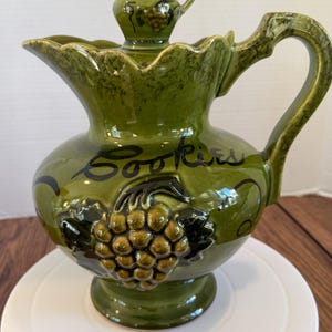 May include: Green ceramic cookie jar with a matching lid and small pitcher on top. The jar has a bulbous shape, a handle, and a spout. The word "Cookies" is written on the front, and a grape cluster design is featured.
