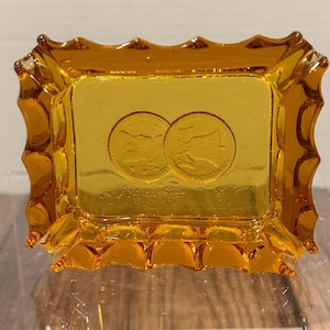 May include: Amber-colored glass tray with a rectangular base and scalloped edges. Two circular coin impressions are visible in the center of the tray. The tray is made of transparent glass.