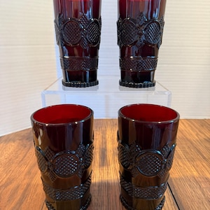 May include: Four ruby red drinking glasses. The glasses have a textured pattern with a repeating design of circles and diamond shapes. The glasses are cylindrical with a slightly flared top and a solid base. The glasses are displayed on a clear surface and a wooden surface.