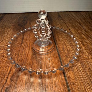 May include: Clear glass cake stand with a beaded edge and a decorative center. The stand has a round base and a central design with a spherical top. The stand is on a wooden surface.