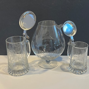 Op de afbeelding: Een set van drie heldere gegraveerde drinkbekers van glas met zilveren deksels. De bekers hebben een bloemmotief en omvatten een kan en twee bekers.