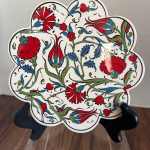 May include: A white, scalloped-edge ceramic tile with a floral design. The design features red, blue, and green flowers and leaves on a white background. The tile is displayed on a black stand.