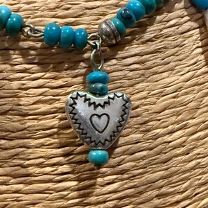 May include: A turquoise beaded necklace with a silver heart pendant. The heart pendant has a scalloped edge and a small heart cutout in the center. The necklace is on a woven background.
