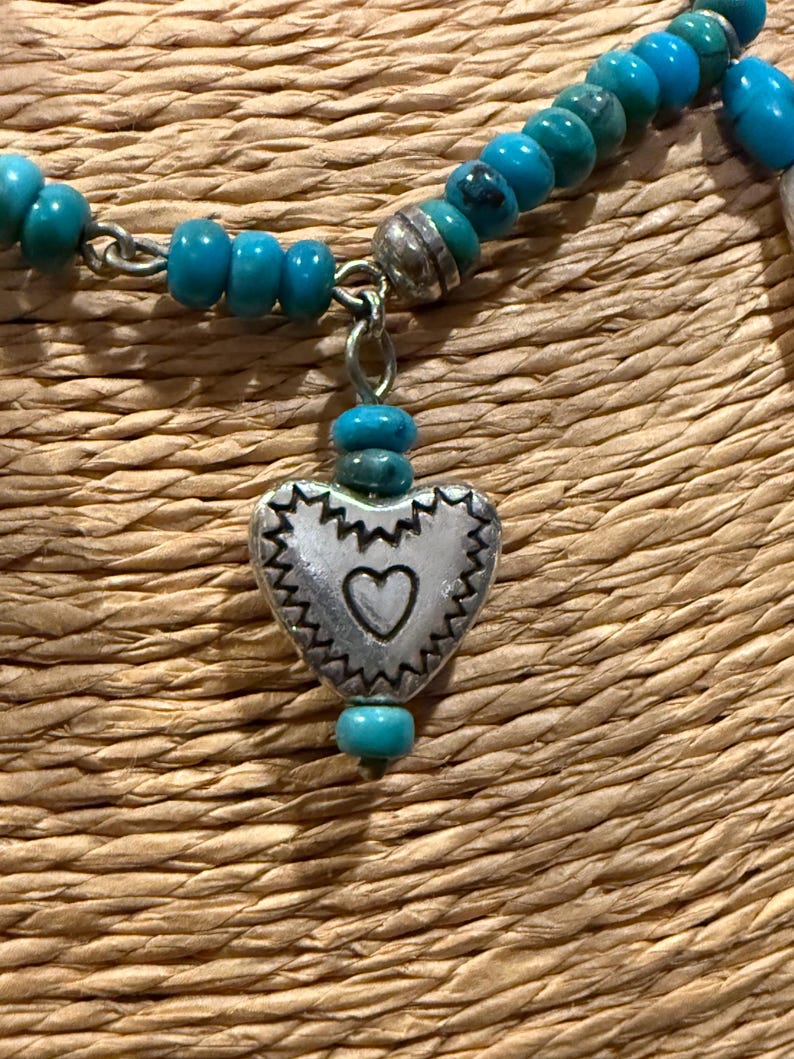 May include: A turquoise beaded necklace with a silver heart pendant. The heart pendant has a scalloped edge and a small heart cutout in the center. The necklace is on a woven background.