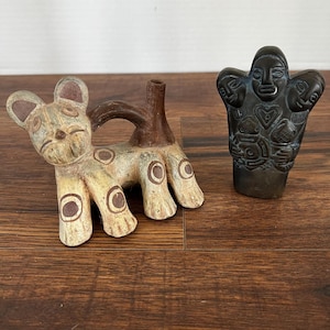 May include: Two ceramic figurines. One is a light-colored cat-like creature with brown spots and ears. The other is a dark-colored, stylized figure with multiple faces and intricate patterns. Both are on a wooden surface.