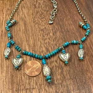 May include: A silver-toned necklace with turquoise beads and heart-shaped pendants. The necklace has a chain and a clasp. A penny is next to the necklace for size reference. The necklace is on a wooden surface.