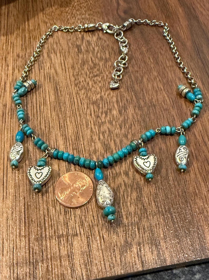 May include: A silver-toned necklace with turquoise beads and heart-shaped pendants. The necklace has a chain and a clasp. A penny is next to the necklace for size reference. The necklace is on a wooden surface.